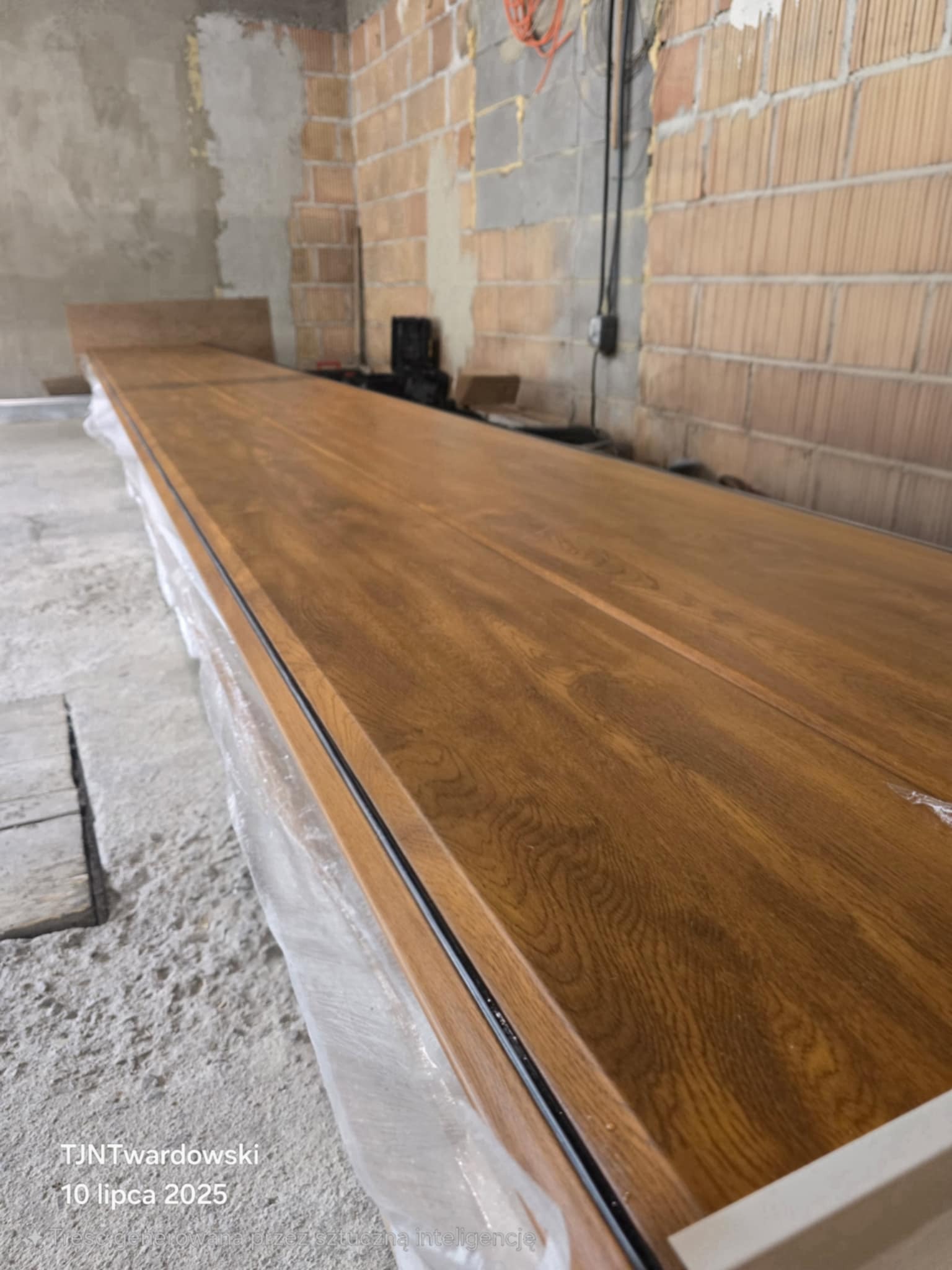 Długa, drewniana brama garażowa w trakcie montażu, zabezpieczona folią, na tle surowej ściany z cegły i betonu. Widoczne elementy konstrukcyjne.