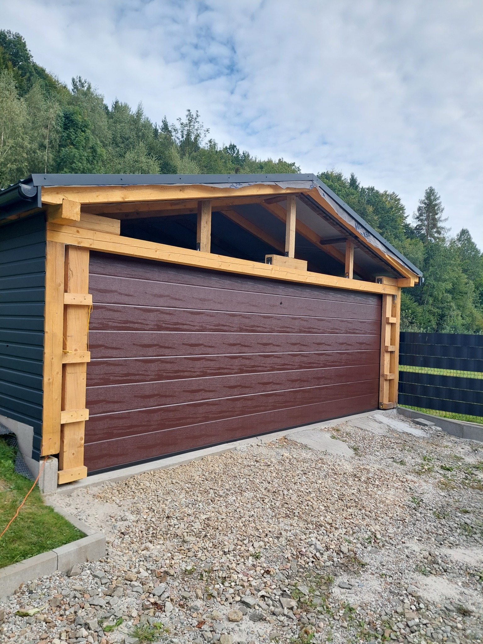 Brązowa brama garażowa segmentowa w trakcie montażu, widoczne drewniane elementy konstrukcyjne i otoczenie posesji z zielenią w tle.