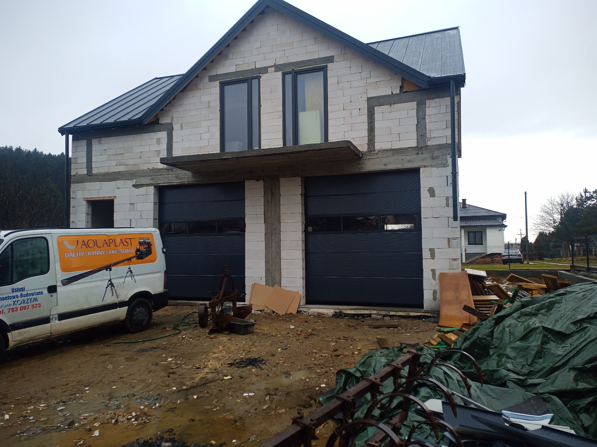 Dom w trakcie budowy z zamontowanymi, grafitowymi bramami garażowymi segmentowymi. Na pierwszym planie teren budowy i samochód dostawczy.