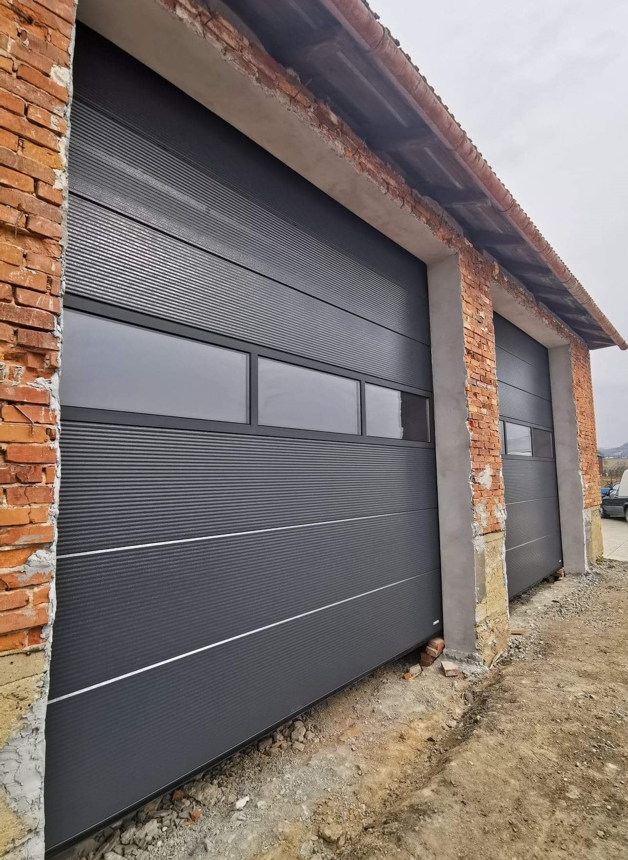 Dwie szare bramy segmentowe z oknami, zamontowane w surowych, ceglanych murach budynku. Widoczne detale wykończenia i surowy stan otoczenia.