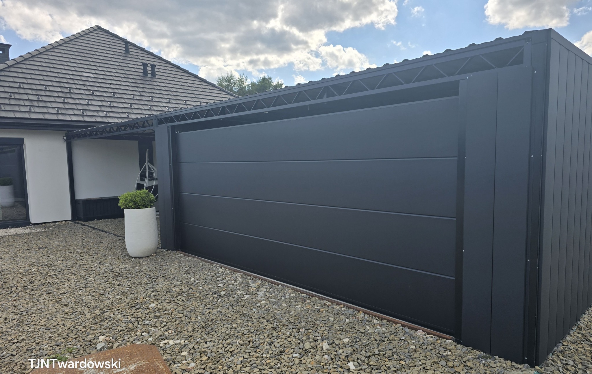 Nowoczesna, grafitowa brama garażowa segmentowa, z widoczną konstrukcją nośną, na tle domu z szarą dachówką i żwirowym podłożem. Minimalistyczny design.