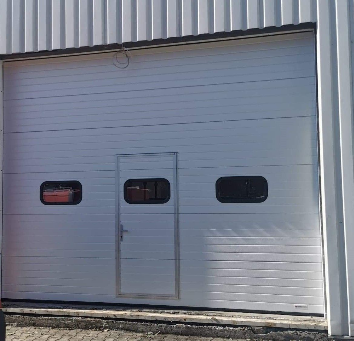 Biała segmentowa brama garażowa z trzema okienkami i drzwiami wejściowymi, zamontowana w hali o metalowej elewacji. Widoczne narzędzia wewnątrz przez okienka.