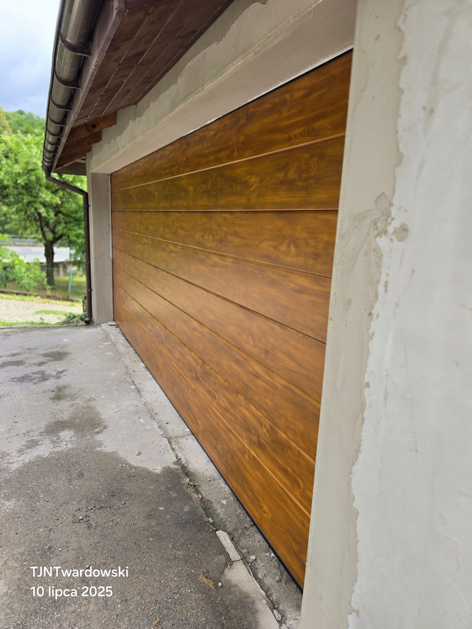 Zewnętrzny widok na segmentową bramę garażową w odcieniach brązu, z widocznymi poziomymi panelami. Świeżo otynkowana ściana garażu w tle. Ujęcie z dołu.