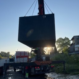 Beton-Tank - Żuraw samochodowy podnosi betonowy zbiornik na tle wschodzącego słońca. Widoczne łańcuchy i fragmenty ciężarówki. Teren błotnisty, zieleń w tle.