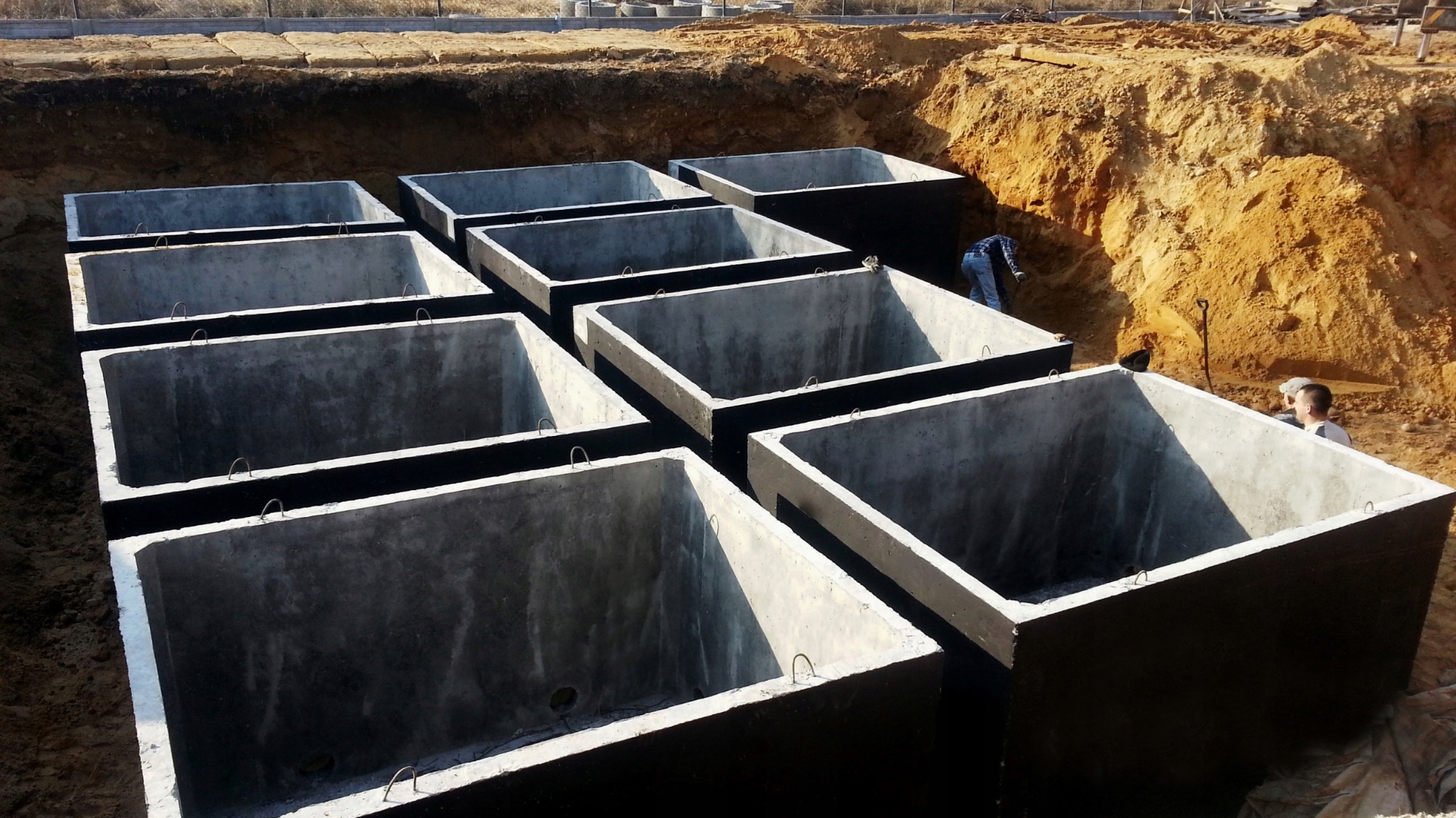 Szamba betonowe w Kartuzach, przygotowane do montażu w wykopie. Widoczne stalowe uchwyty i uszczelnienie. Praca w toku, teren budowy.