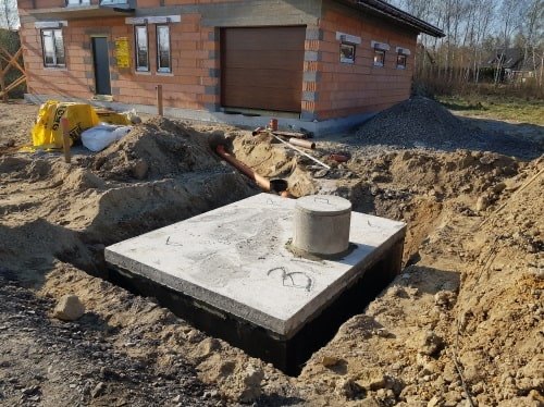 Betonowy zbiornik na szambo, częściowo zakopany w ziemi, z widoczną pokrywą. W tle budynek w trakcie budowy i hałda żwiru.