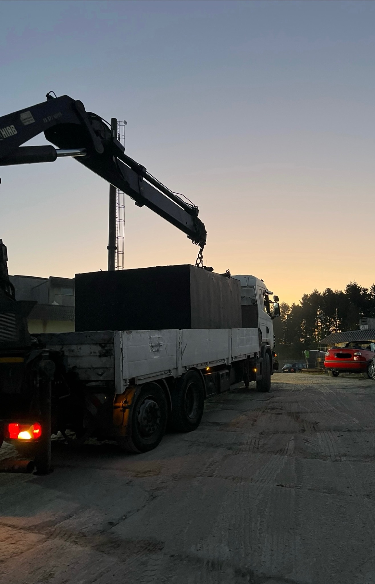 Transport szamba betonowego dźwigiem na ciężarówce o świcie. Widoczne drzewa w tle i czerwony samochód. Szary betonowy zbiornik na platformie.