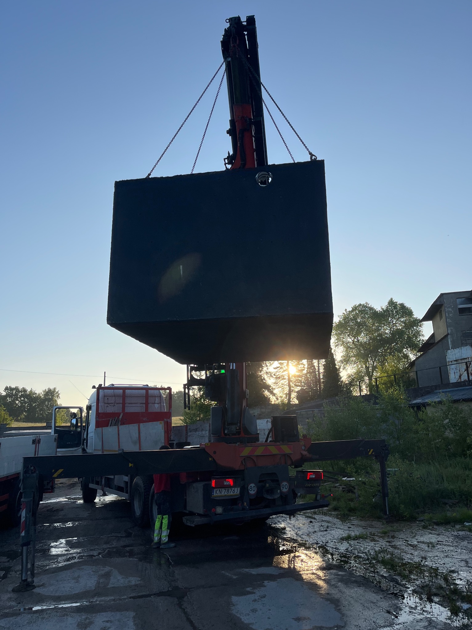 Żuraw samochodowy podnosi betonowy zbiornik na tle wschodzącego słońca. Widoczne łańcuchy i fragmenty ciężarówki. Teren błotnisty, zieleń w tle.