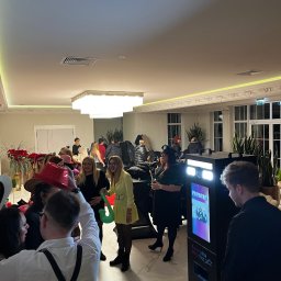 Fabryka Imprez - Uczestnicy imprezy pozują do zdjęcia w fotobudce 'Fabryka Imprez' w eleganckim wnętrzu. Widoczne rekwizyty i uśmiechnięte osoby.