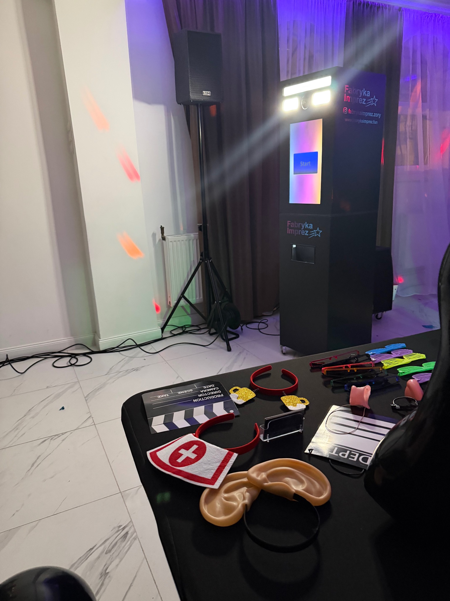 Fotobudka 'Fabryka Imprez' z rekwizytami: klaps filmowy, okulary, uszy elfa i opaska pielęgniarki. W tle kolumna głośnikowa i fioletowe oświetlenie.