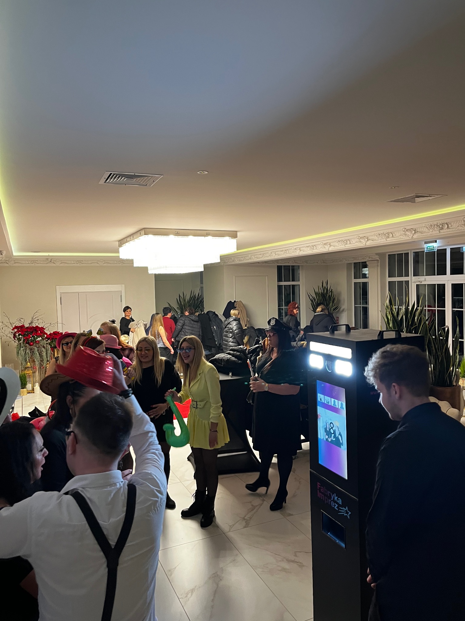 Uczestnicy imprezy pozują do zdjęcia w fotobudce 'Fabryka Imprez' w eleganckim wnętrzu. Widoczne rekwizyty i uśmiechnięte osoby.