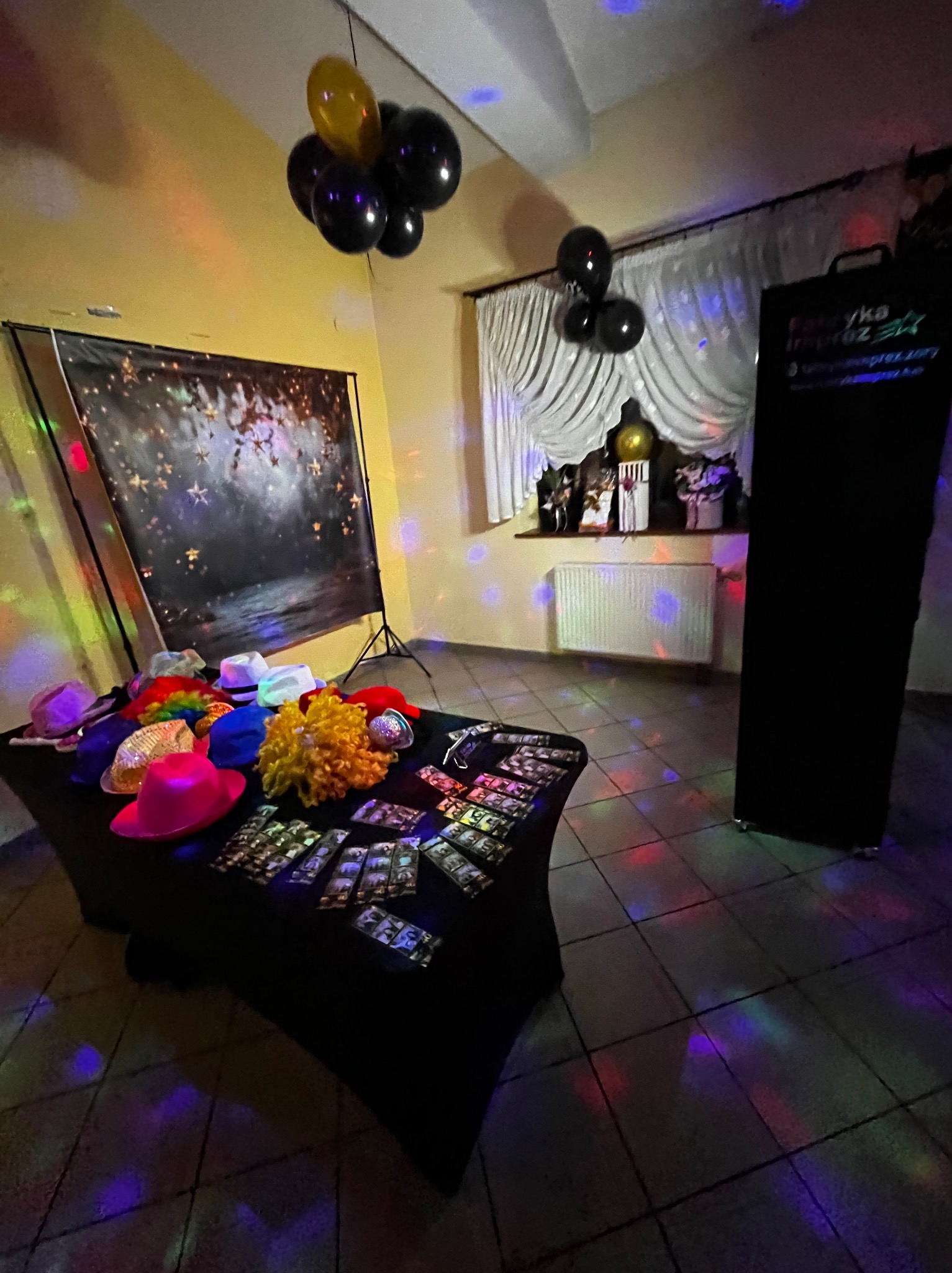 Stanowisko fotobudki z rekwizytami: kolorowe kapelusze, peruki, tło z gwiazdami, balony. Przygotowane do sesji zdjęciowej na imprezie, oświetlone kolorowymi światłami.