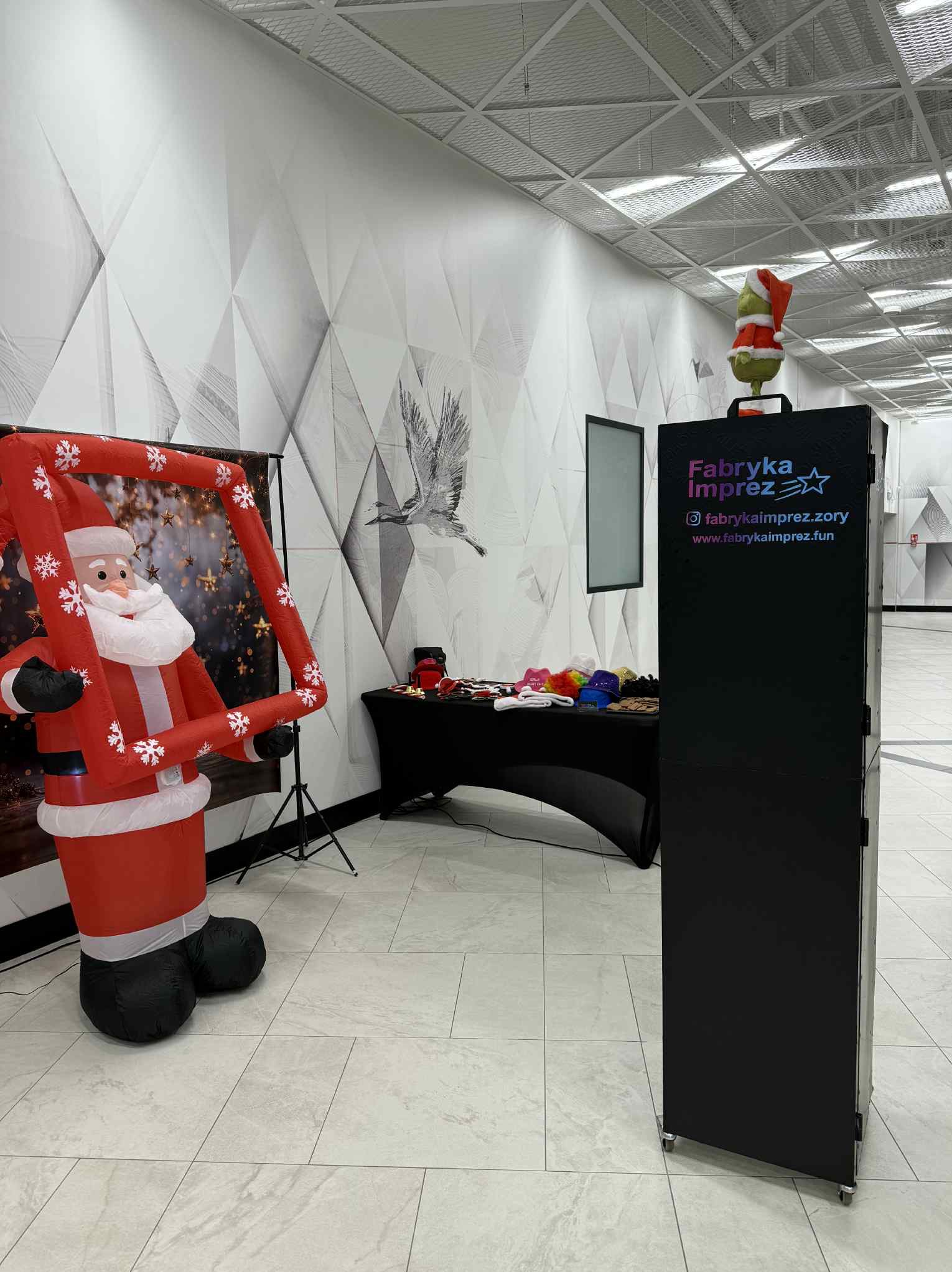 Inflatowany Mikołaj z ramką, fotobudka z logo firmy, stół z rekwizytami na tle wzorzystej ściany. Gotowość do świątecznej sesji zdjęciowej.