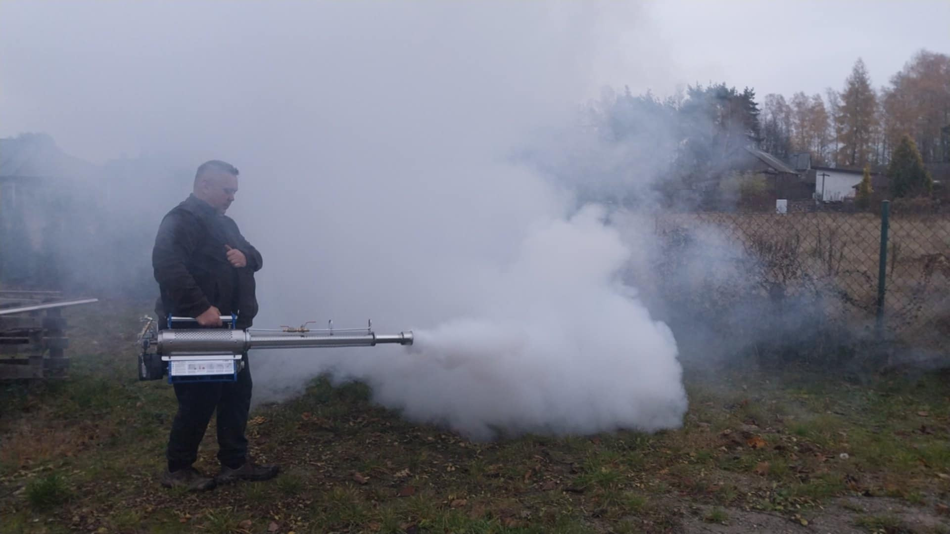 Osoba w czarnej kurtce obsługuje zamgławiacz termiczny, generujący gęstą mgłę na zewnątrz, w tle widoczny dom i drzewa.