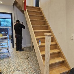 R-BAU USŁUGI REMONTOWO-BUDOWLANE ROBERT PŁOTKA - Montaż drewnianych schodów w jasnym wnętrzu. Widoczny pracownik podczas pracy, fragment wzorzystej podłogi i butelka kleju na schodach.