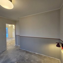 R-BAU USŁUGI REMONTOWO-BUDOWLANE ROBERT PŁOTKA - Wnętrze w trakcie malowania: ściany dwukolorowe (szary dół, beżowa góra) oddzielone brązowym paskiem, lampka ścienna, surowa podłoga, widok na kolejne pomieszczenie.