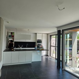 R-BAU USŁUGI REMONTOWO-BUDOWLANE ROBERT PŁOTKA - Nowoczesna kuchnia z czarną podłogą i białymi szafkami, widok na ogród przez przesuwne drzwi. W tle drabina oparta o ścianę, sugerująca prace wykończeniowe.