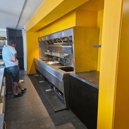 R-BAU USŁUGI REMONTOWO-BUDOWLANE ROBERT PŁOTKA - Żółta kuchnia komercyjna z metalową zabudową i czarnymi szafkami, widok z boku, osoba w tle. Nietypowe, jaskrawe wykończenie wnętrza lokalu gastronomicznego.