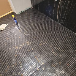 R-BAU USŁUGI REMONTOWO-BUDOWLANE ROBERT PŁOTKA - Ukończona podłoga z mozaiki w łazience, z ciemnymi płytkami i złotymi akcentami. Widoczne narzędzia i czyszczące środki. Ściana wykończona czarnym marmurem z żyłami.