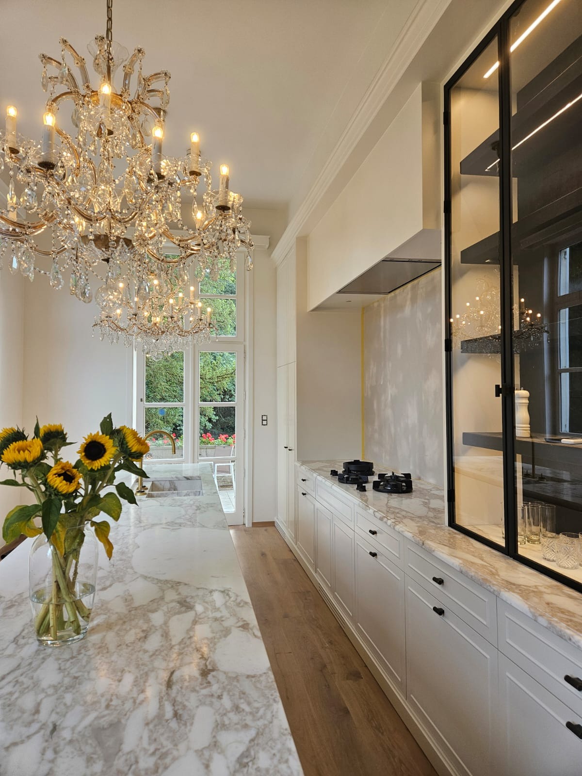 Elegancka kuchnia z marmurowym blatem, kryształowym żyrandolem i bukietem słoneczników. Widoczne białe szafki i przeszklona witryna. Ściana przygotowana do malowania.