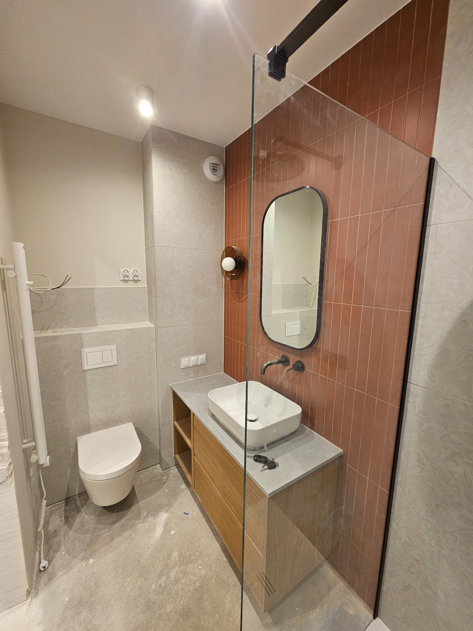 Nowoczesna łazienka z umywalką nablatową, lustrem w czarnej ramie i płytkami w odcieniach terakoty. Widoczny fragment kabiny prysznicowej i wisząca toaleta.