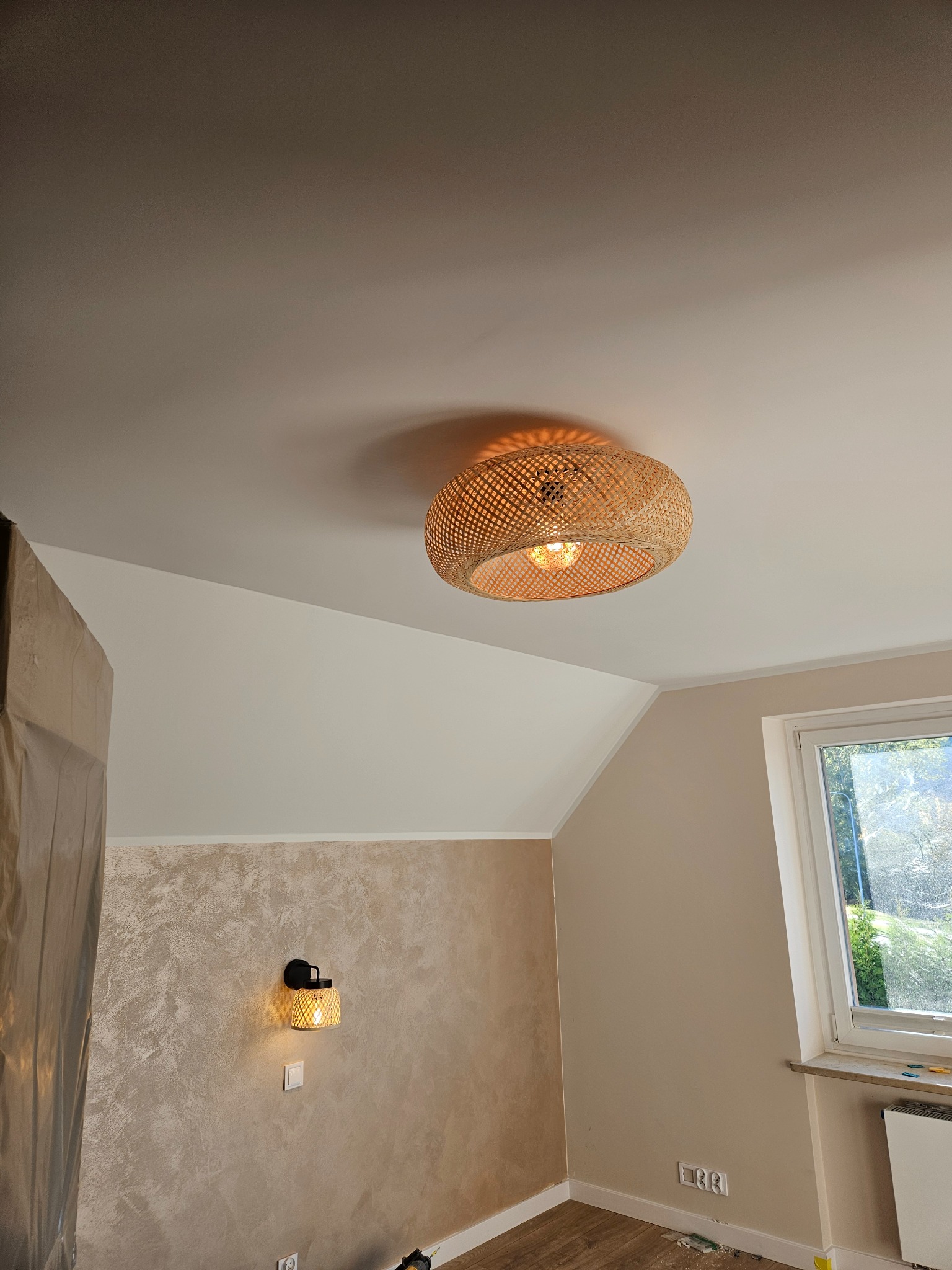 Wykończone wnętrze pokoju z beżowymi ścianami, dekoracyjną strukturą na jednej ścianie, oświetleniem w stylu boho i oknem z widokiem na zieleń.