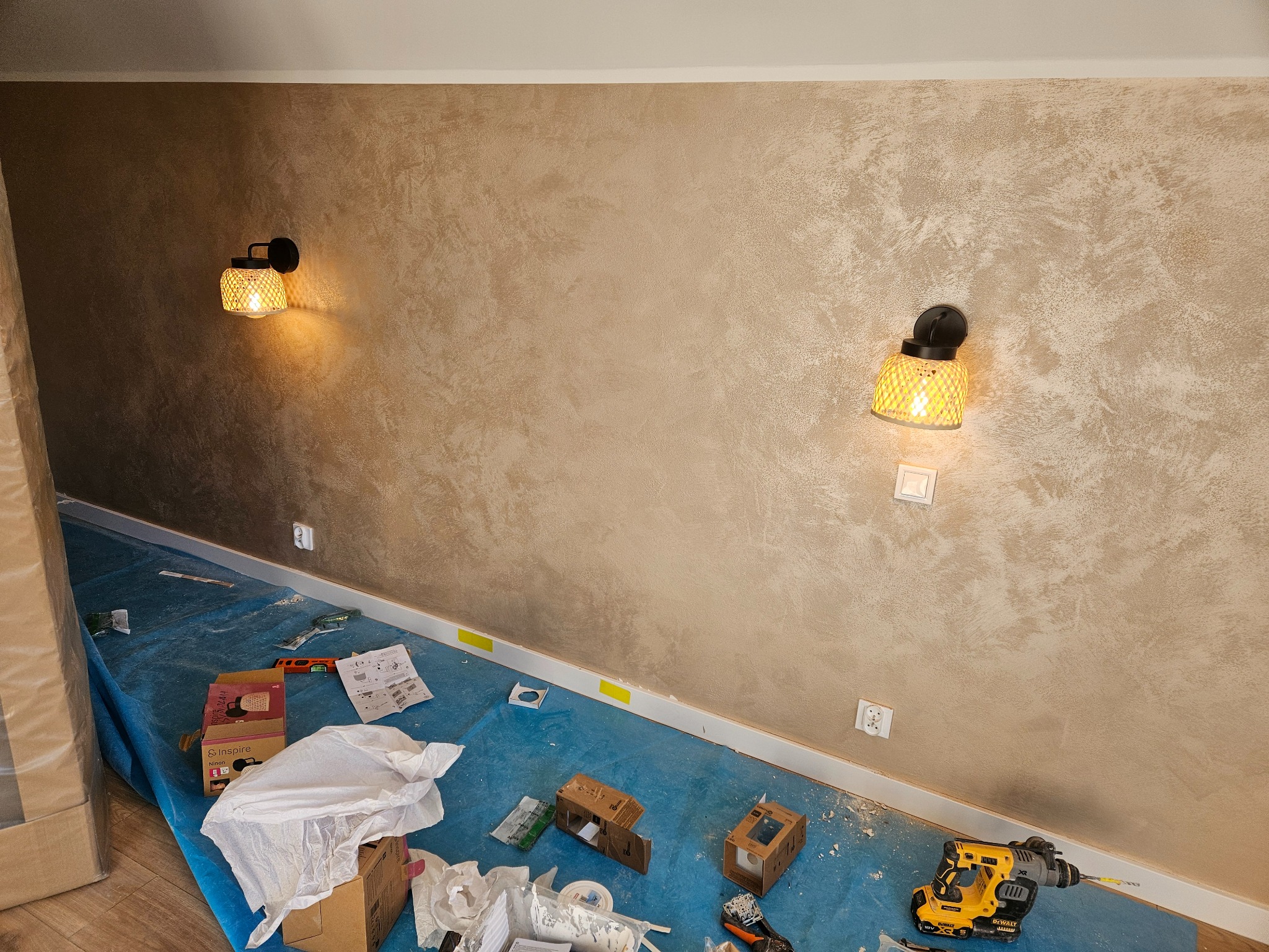 Ściana z dekoracyjną strukturą w odcieniach beżu, oświetlona dwoma kinkietami z ażurowymi kloszami. Podłoga zabezpieczona folią, widoczne narzędzia i materiały.