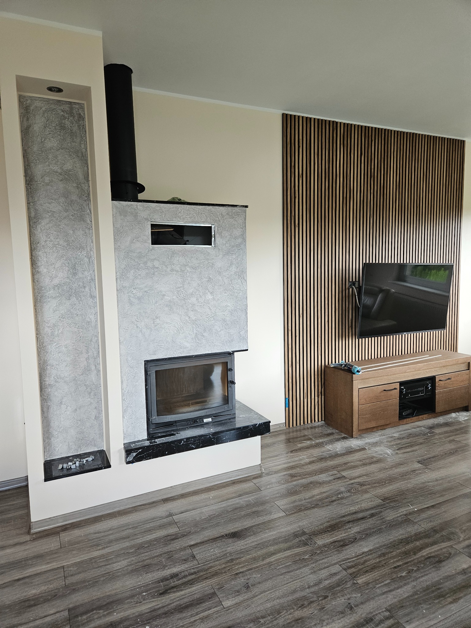 Nowoczesny salon z kominkiem obłożonym szarym tynkiem strukturalnym, drewnianą ścianą lamelową i wiszącym telewizorem. Podłoga wyłożona panelami imitującymi drewno.