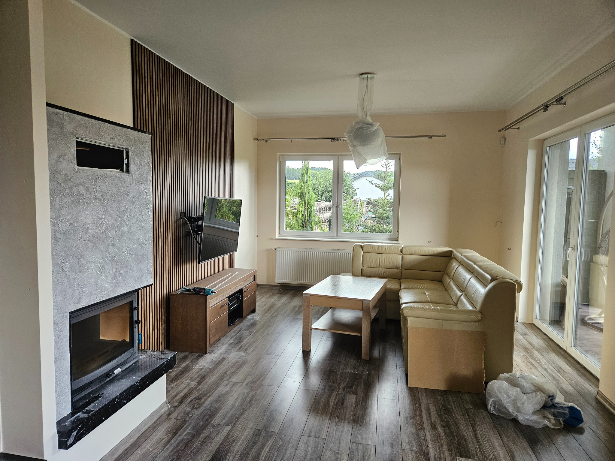 Salon po remoncie w Luzinie: drewniane lamele na ścianie, kominek z szarą strukturą, podłoga z paneli. Widoczne meble i okno z widokiem na zieleń.