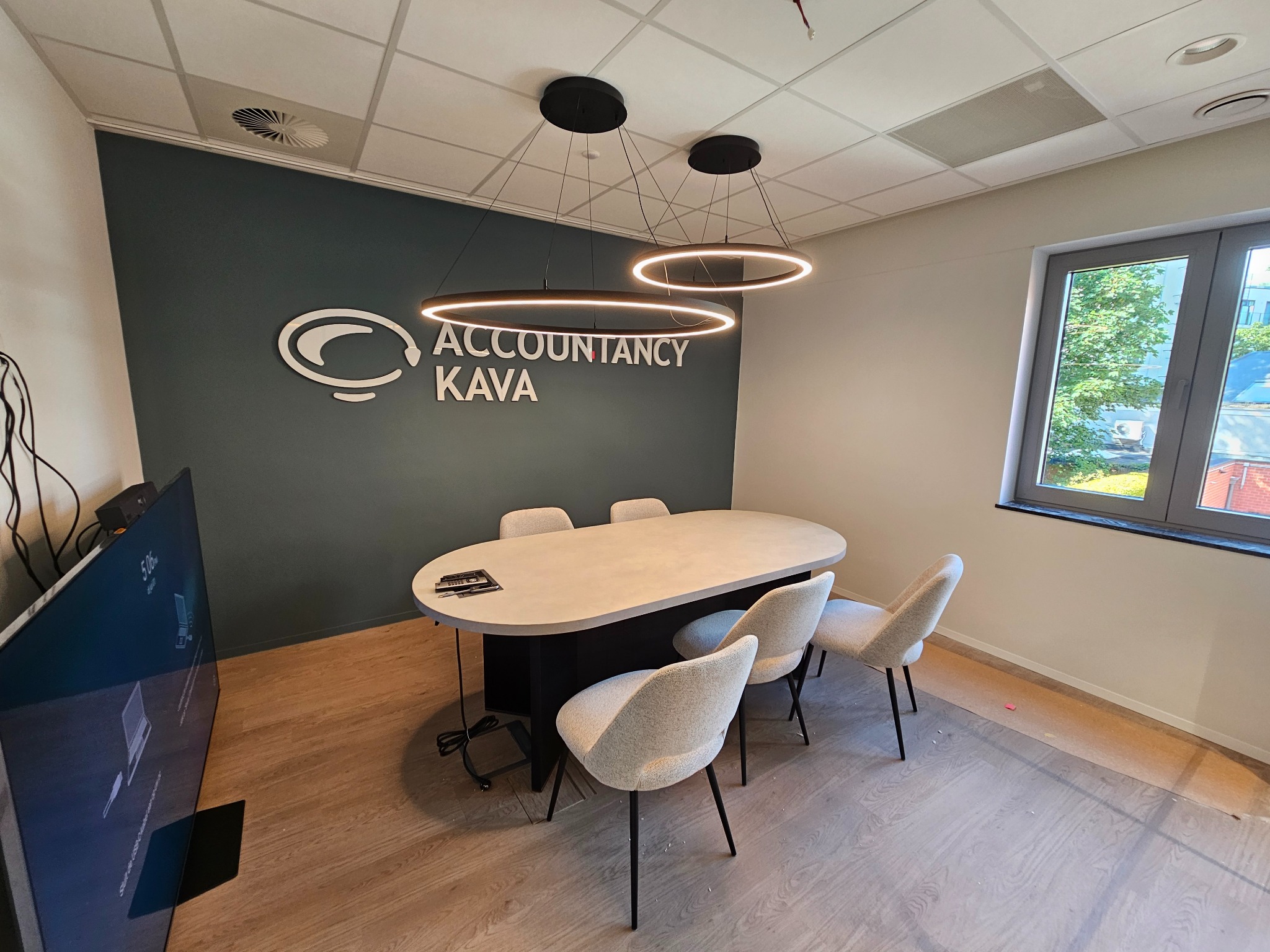 Nowoczesne biuro z owalnym stołem konferencyjnym, krzesłami i designerskimi lampami. Na ścianie logo firmy Accountancy Kava. Jasne drewniane podłogi i duże okno.
