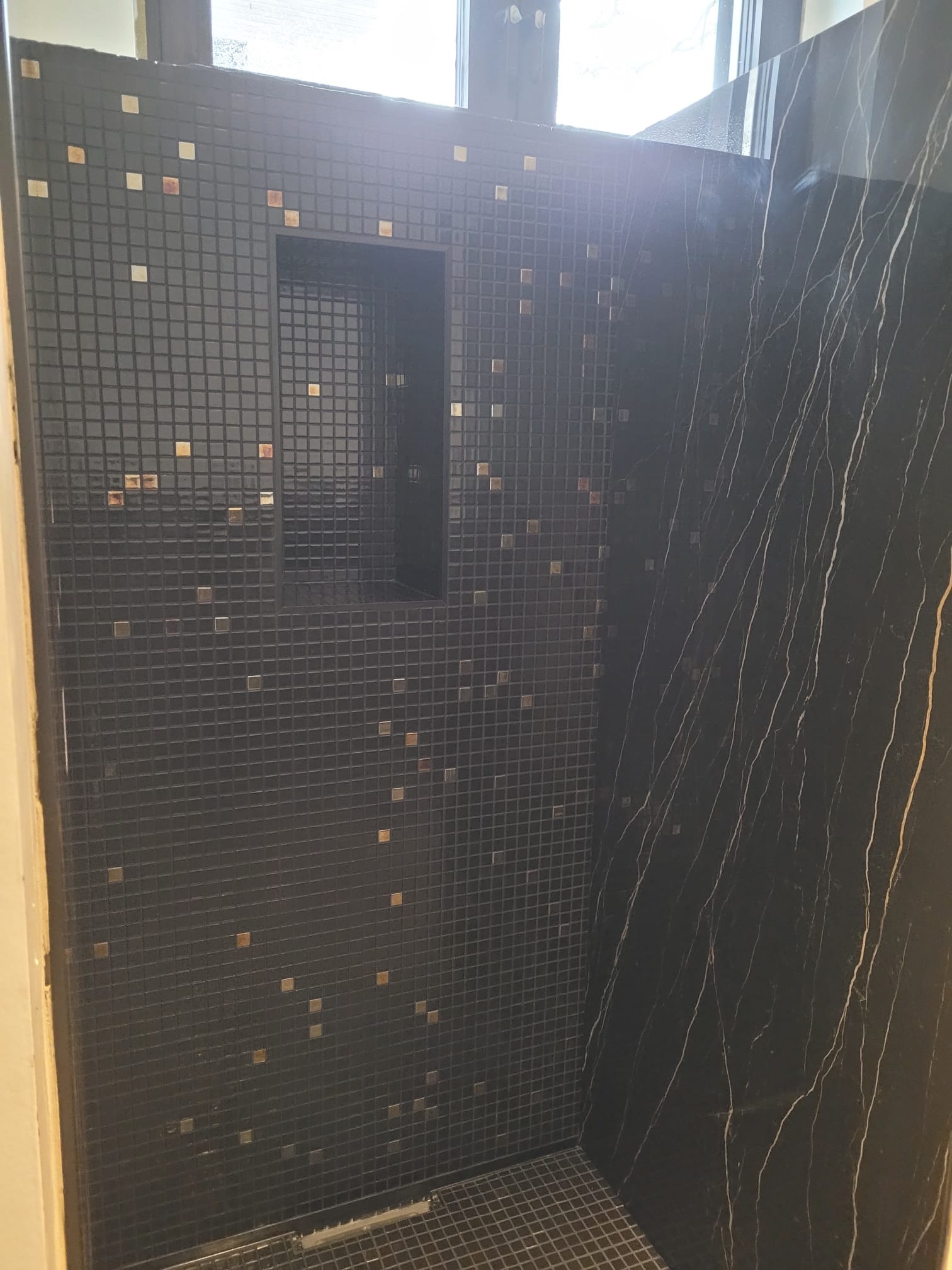 Wykończona kabina prysznicowa z czarną mozaiką i wnęką, akcentowana złotymi elementami. Obok ściana z czarnego marmuru z białymi żyłami.