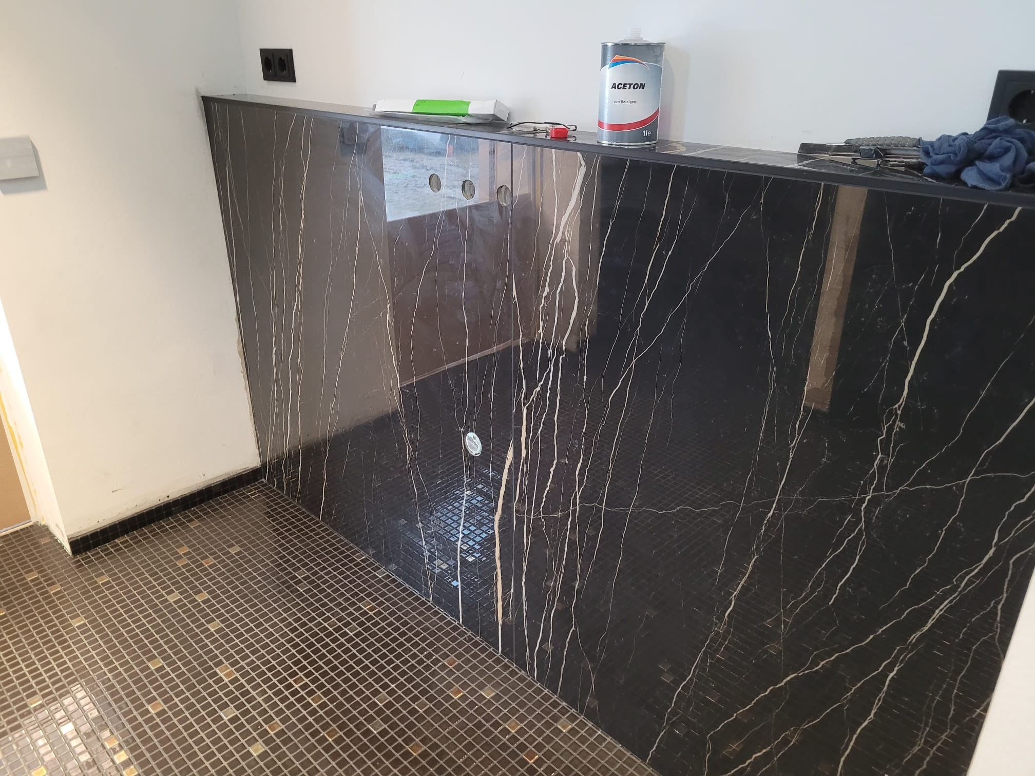 Wykończona ściana i podłoga w łazience z czarnymi płytkami imitującymi marmur z białymi żyłami. Widoczne narzędzia i chemia na blacie.