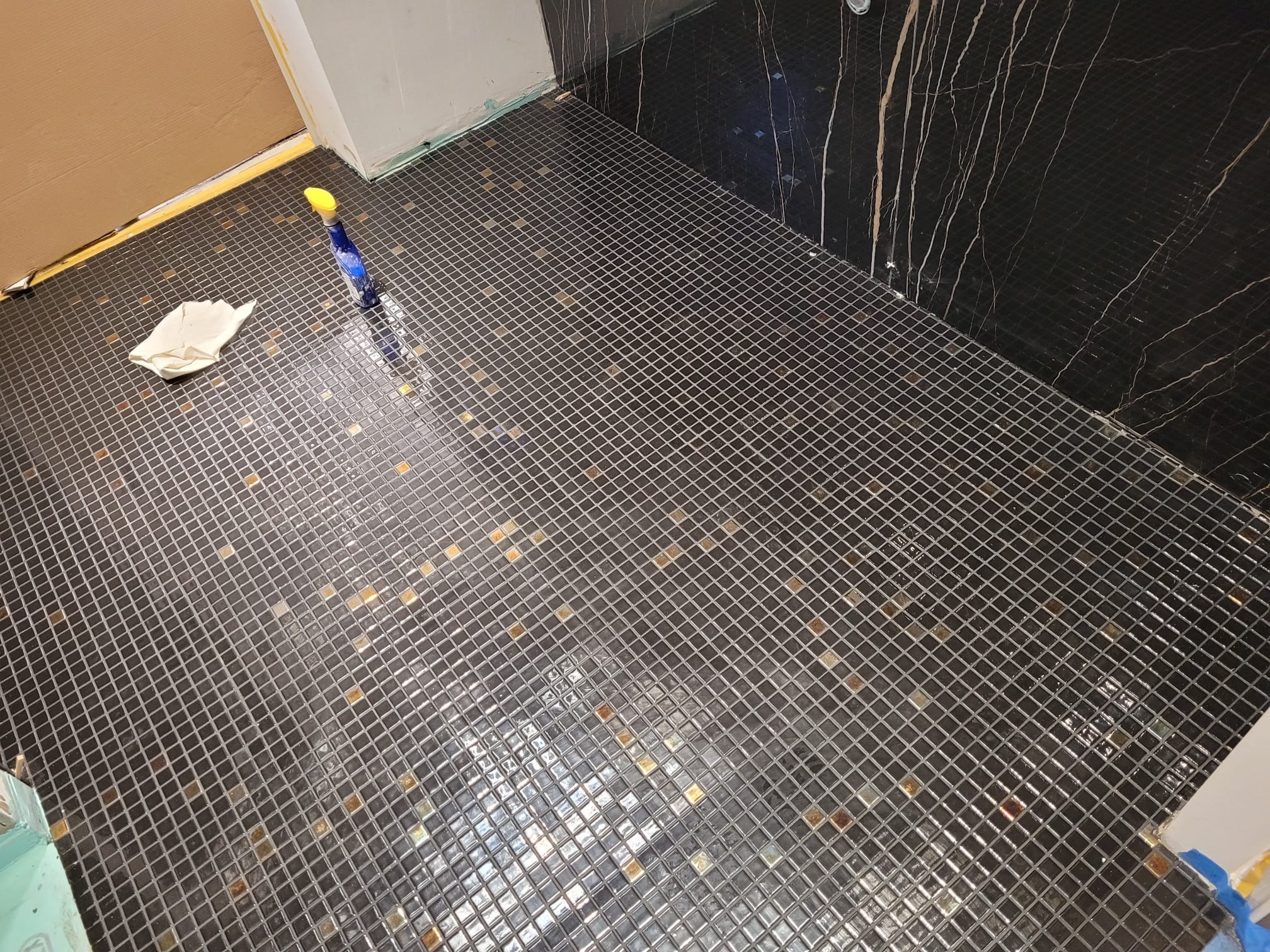 Ukończona podłoga z mozaiki w łazience, z ciemnymi płytkami i złotymi akcentami. Widoczne narzędzia i czyszczące środki. Ściana wykończona czarnym marmurem z żyłami.