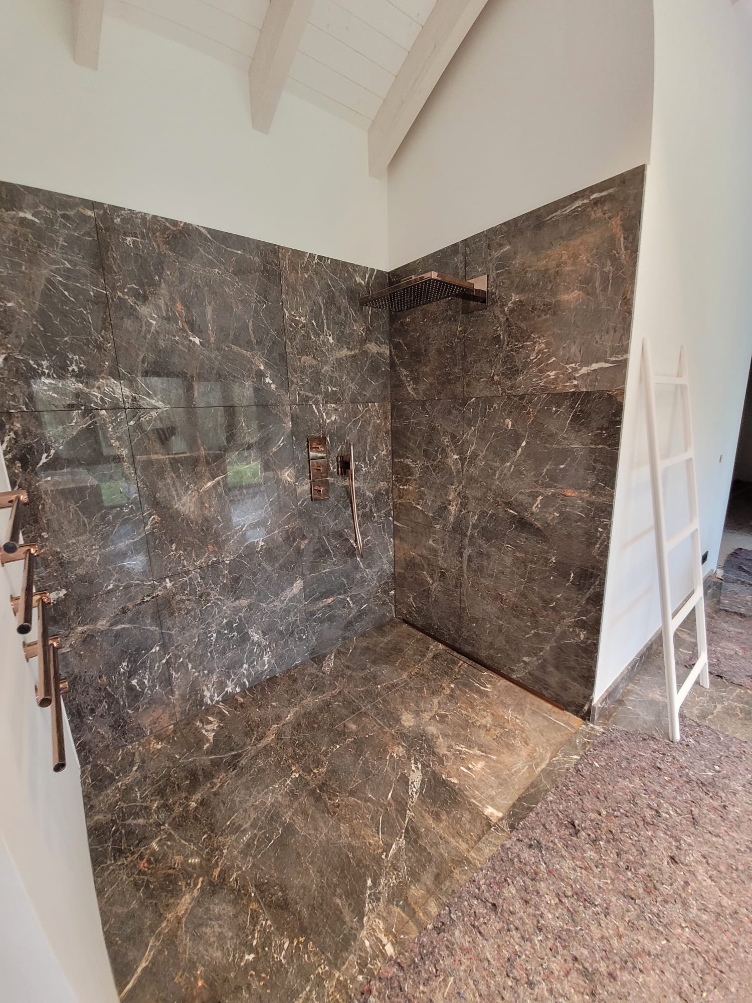 Wykończona kabina prysznicowa z ciemnymi płytkami imitującymi marmur, z miedzianą armaturą i białą drabinką dekoracyjną. Widoczne skosy sufitu.