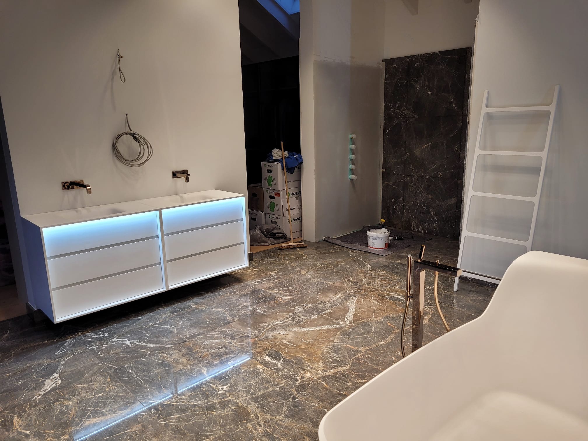 Łazienka w trakcie wykańczania: marmurowa podłoga, podświetlana szafka pod umywalkę, drabinka dekoracyjna i fragment wanny. Widoczne elementy instalacji.