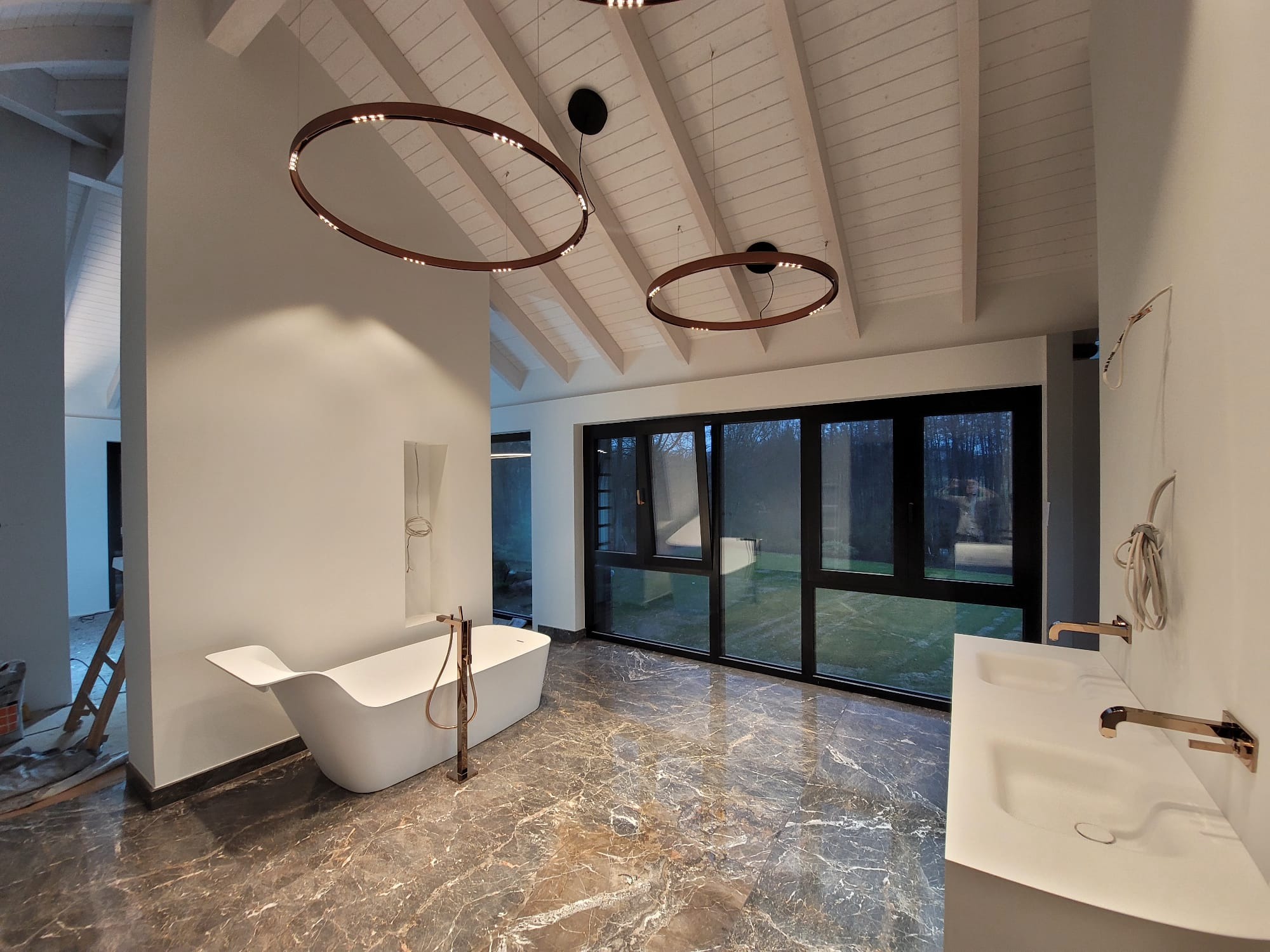 Nowoczesna łazienka z marmurową podłogą, wolnostojącą wanną i podwójną umywalką. Okrągłe lampy sufitowe i duże okno z widokiem na zieleń.