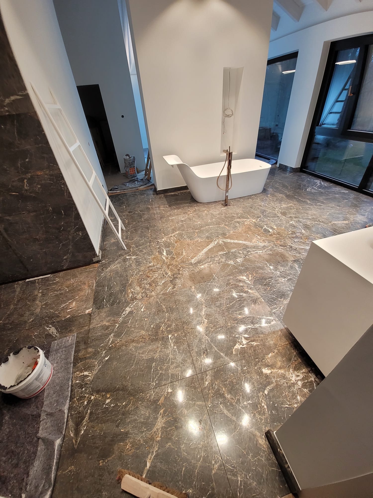 Luksusowa łazienka w trakcie wykończenia: marmurowe płytki podłogowe, wolnostojąca wanna, duże okna. Widoczne narzędzia glazurnicze i drabina.
