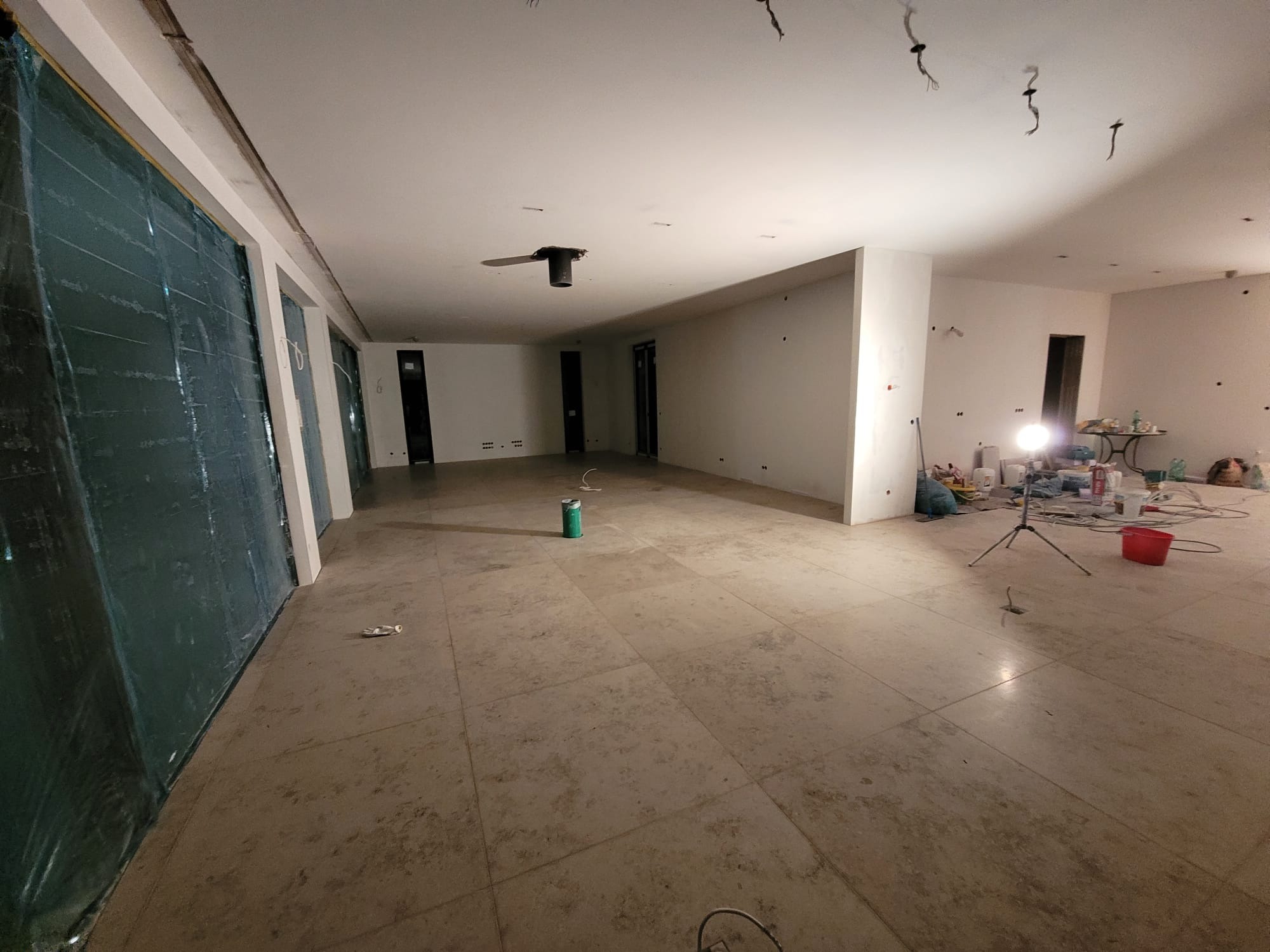 Przestronne wnętrze w trakcie remontu: zabezpieczone folią okna, surowa podłoga z płytek, ściany przygotowane do malowania, widoczne kable i narzędzia malarskie.