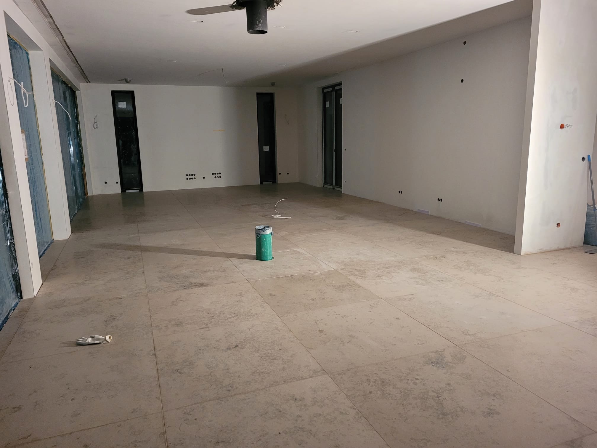 Przestronne wnętrze w trakcie prac wykończeniowych: jasne płytki podłogowe, ściany przygotowane do malowania, widoczne puszki elektryczne i pojedyncza puszka z farbą.