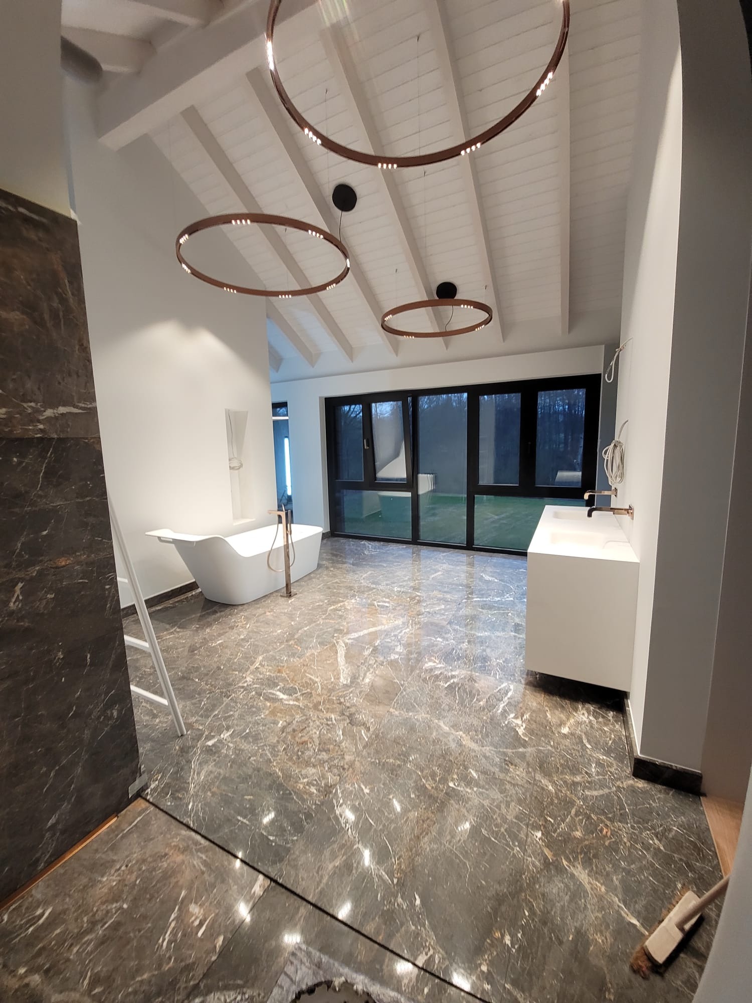 Nowoczesna łazienka z marmurową podłogą, wolnostojącą wanną i okrągłymi lampami sufitowymi. Duże okno z widokiem na zieleń. Minimalistyczna umywalka.