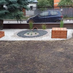 Daw Garden - Ogród po aranżacji: drewniane skrzynie, ozdobny okrąg z kamieni, trawa w trakcie wzrostu. W tle czarny płot i dom.