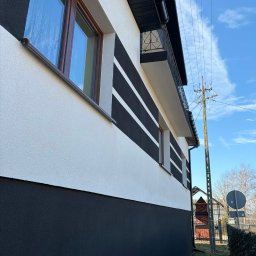 Cichy Kąt USŁUGI REMONTOWO - BUDOWLANE Maciej Dobiesz - Elewacja domu po malowaniu w biało-czarne pasy, z widocznym balkonem i oknami z brązowymi ramami. Słup elektryczny w tle.