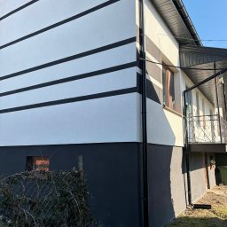 Cichy Kąt USŁUGI REMONTOWO - BUDOWLANE Maciej Dobiesz - Elewacja domu z poziomymi, czarnymi pasami na białym tle i ciemnoszarym cokołem. Widoczny fragment balkonu i rynny. Ujęcie z dołu, w słoneczny dzień.