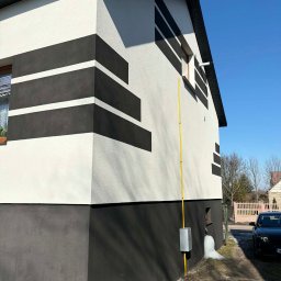 Cichy Kąt USŁUGI REMONTOWO - BUDOWLANE Maciej Dobiesz - Dwukolorowa elewacja domu z poziomymi pasami, widok z boku, błękitne niebo w tle. Żółta rura gazowa przy ścianie. Nowoczesny design.