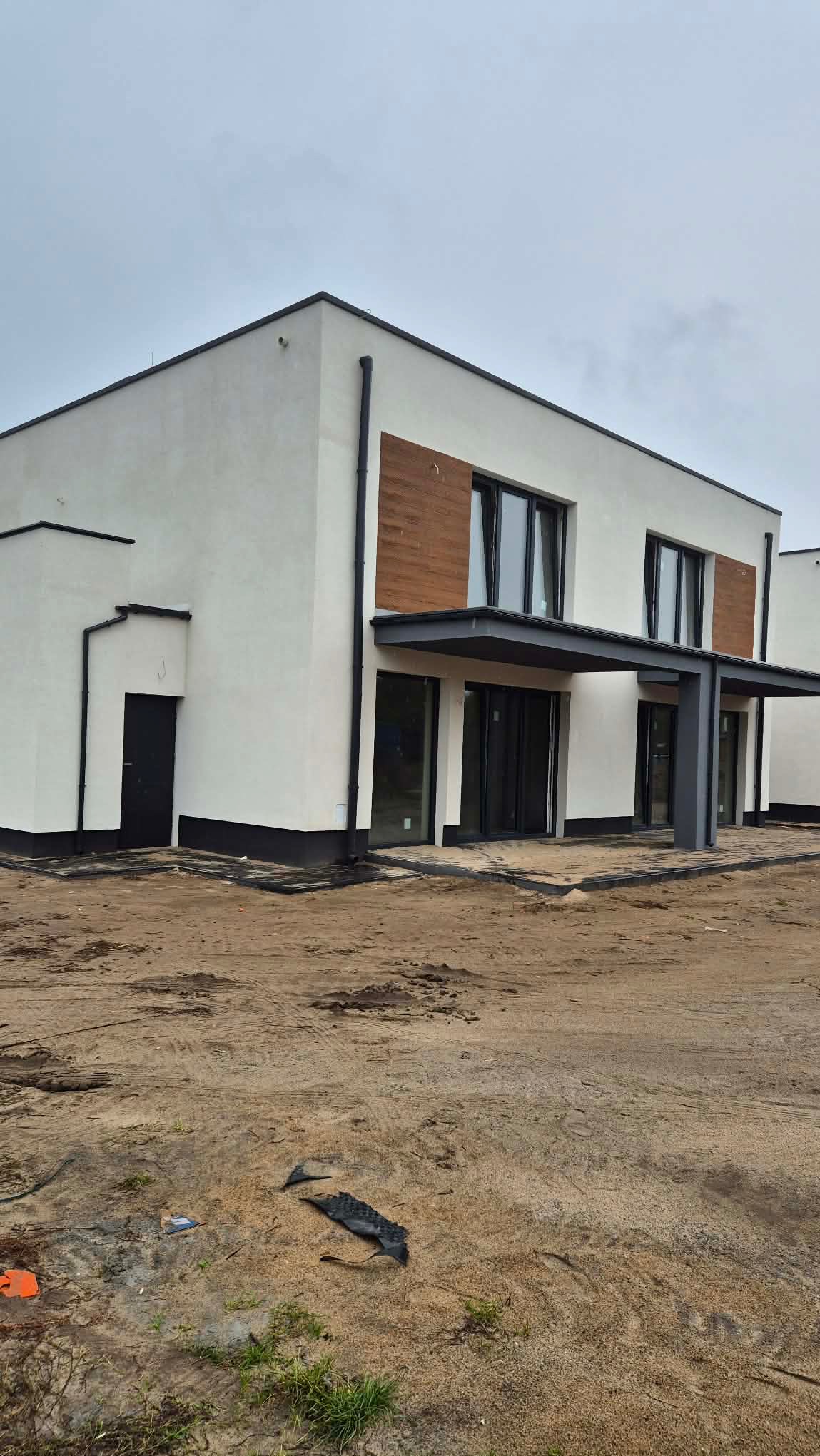 Nowoczesny, dwukondygnacyjny dom z białą elewacją, drewnianymi akcentami i czarnymi ramami okiennymi. Widoczne zadaszenia nad wejściami i teren wokół budynku w trakcie prac wykończeniowych.