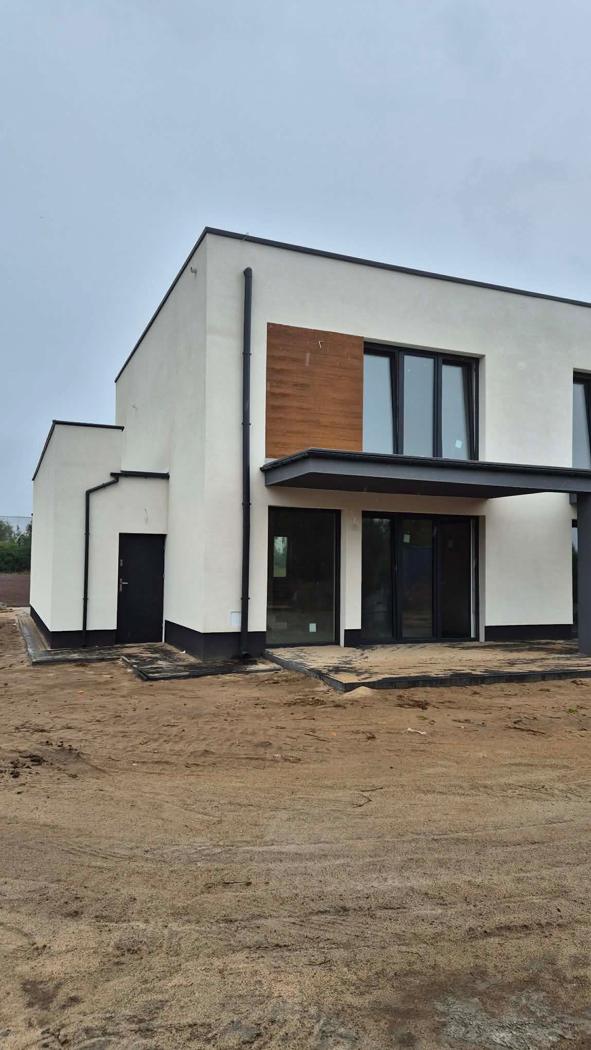 Nowoczesny dom z białą elewacją, czarnymi detalami i drewnianym akcentem. Widoczne okna, drzwi i rynny. Teren wokół domu przygotowany pod zagospodarowanie.