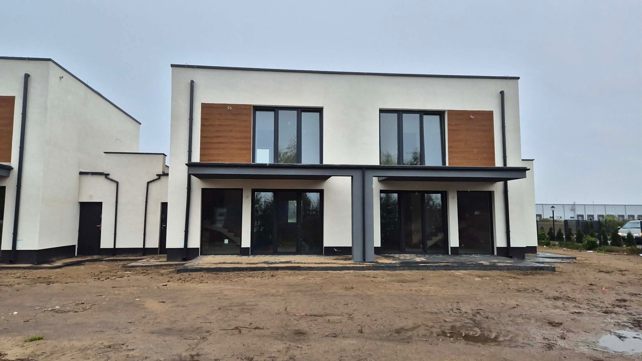 Nowoczesne, dwukondygnacyjne domy bliźniacze z białymi elewacjami i drewnianymi akcentami. Czarne ramy okien i drzwi, minimalistyczny design, surowy krajobraz.