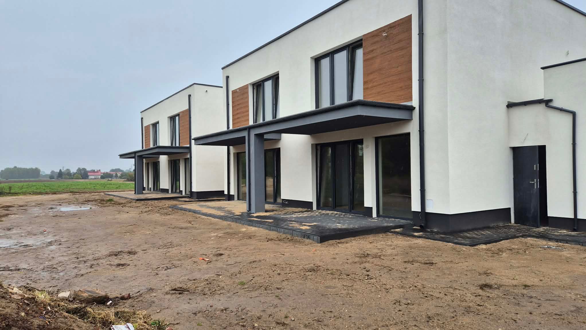 Nowoczesne, szeregowe domy z białymi elewacjami i drewnianymi akcentami, szare zadaszenia nad wejściami, kostka brukowa, widok na pole w tle, pochmurny dzień.