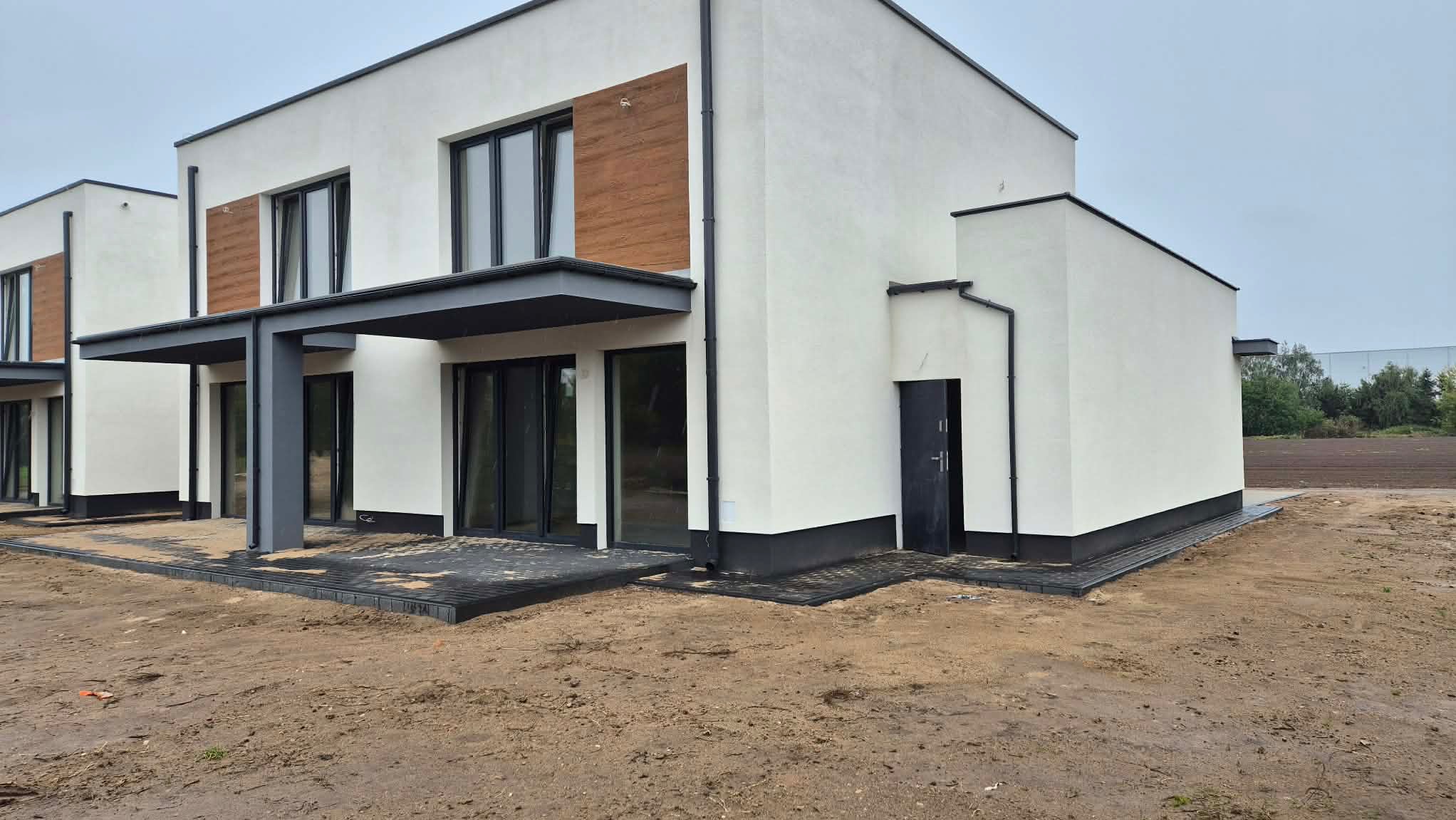 Nowoczesny, biały dom z elementami drewnianymi i szarą podbitką, widoczny z zewnątrz, z niezagospodarowanym terenem wokół. Minimalistyczny design.