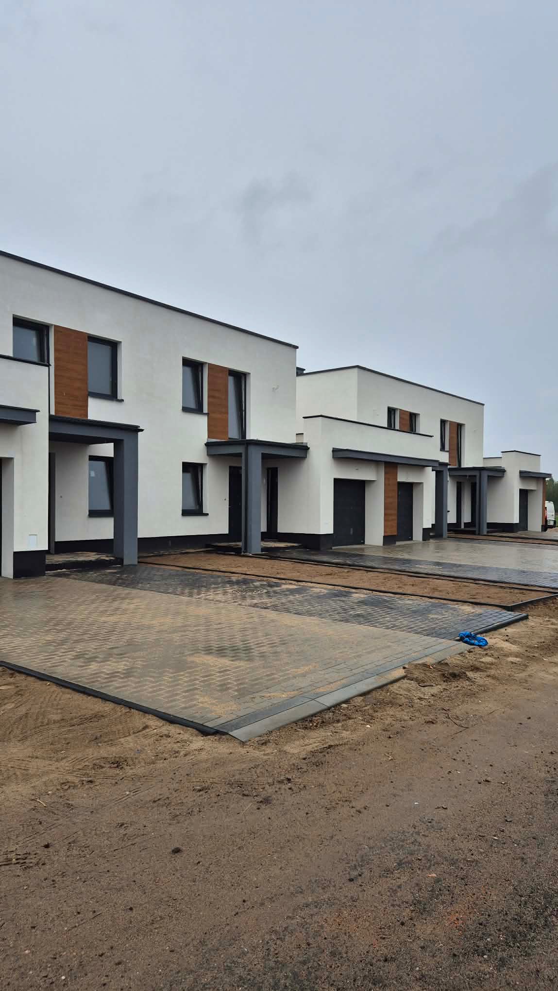 Nowoczesne, szeregowe domy z białymi elewacjami i drewnianymi akcentami, szara kostka brukowa na podjazdach, pochmurne niebo. Widok z poziomu gruntu.